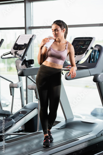 young sportswoman in bra and leggings standing with towel near sports bottle on treadmill