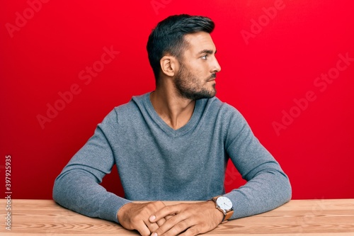 Handsome hispanic man wearing casual style sitting on the table looking to side, relax profile pose with natural face with confident smile.