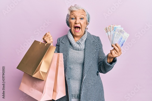 Senior grey-haired woman holding shopping bags and swedish krona banknotes afraid and shocked with surprise and amazed expression, fear and excited face.