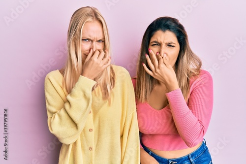 Two friends standing together over pink background smelling something stinky and disgusting, intolerable smell, holding breath with fingers on nose. bad smell