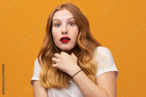 Portrait of the scared girl with wavy redhead, wearing white t-shirt, isolated on yellow background