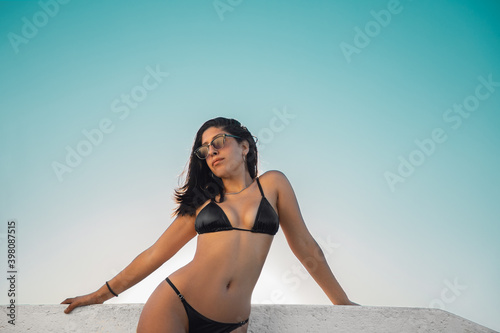 Joven modelo femenina con lentes de sol morocha bronceada en bikini negro posando sobre una baranda blanca en un atardecer de cielo cyan