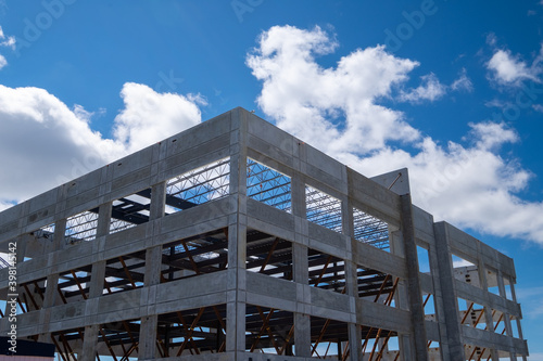 Multiple story precast grey concrete building with steel beams. The industrial structure in the corner of a skyscraper building with prefabricated engineering formwork. The sky is in the background.