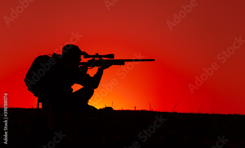 Commando team sniper, army special forces shooter aiming, shooting sniper rifle while sitting on sea or ocean shore during sunset. Coast or border guard soldier observing coastline with optical sight