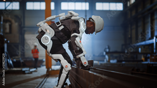 Black African American Engineer is Testing a Futuristic Bionic Exoskeleton and Picking Up Metal Objects in a Heavy Steel Industry Factory. Contractor is Heavy Lifting Steel Parts in a Powered Shell.