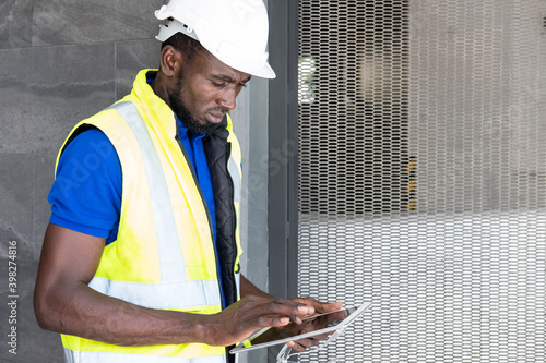 Selective focus at face of Black African foreman at building construction site, wearing protective hat and safety equipment while using digital tablet to record information. Civil engineer working.