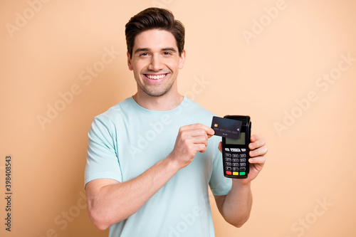 Close-up portrait of nice cheerful guy holding in hand terminal using bank card finance transaction isolated over beige pastel color background