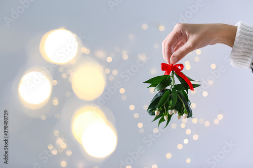 Woman holding mistletoe bunch with red bow against blurred festive lights, closeup and space for text. Traditional Christmas decor