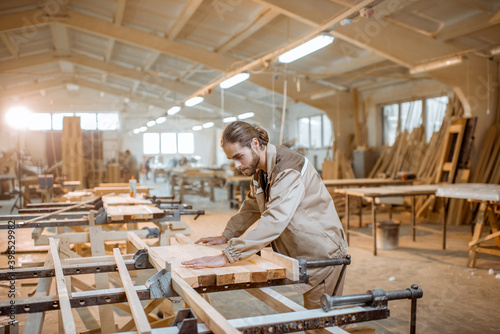 Handsome carpenter in uniform gluing wooden bars with hand pressures at the carpentry manufacturing