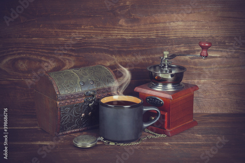 Retro casket, black mug with coffee and steam above it, pocket watch with chain, vintage coffee grinder with handle on brown wooden plank background, tinted.