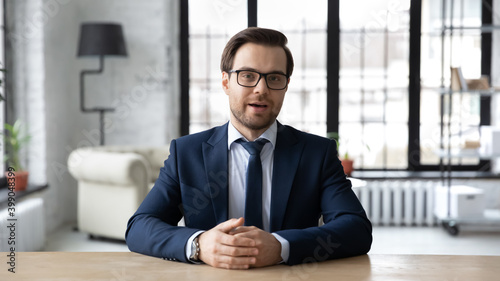 Head shot confident businessman wearing suit and glasses speaking looking at camera, sitting at work desk in office, mentor coach shooting webinar, hr manager holding online interview with candidate