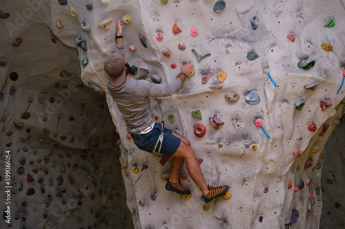 Active sporty man wearing protective face mask and a hat practicing rock climbing on artificial rock in a climbing wall. New normal in extreme sports.