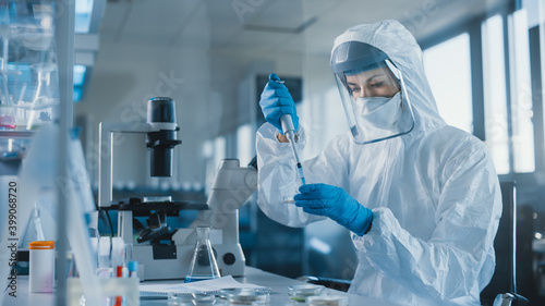 Beautiful Female Medical Scientist Wearing Coverall and Face Mask Using Micro Pipette while Working with Petri Dish. Vaccine, Drugs Research and Development Innovative Laboratory Modern Equipment 