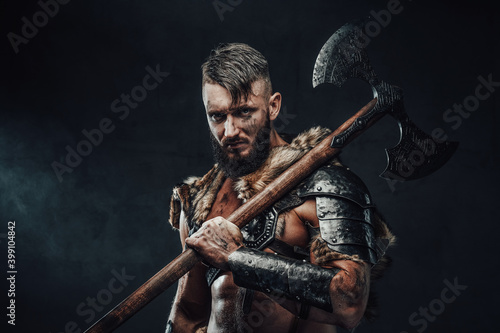 Holding two handed axe on his shoulder scandinavian barbarian in light armour with fur poses in dark background looking at camera.