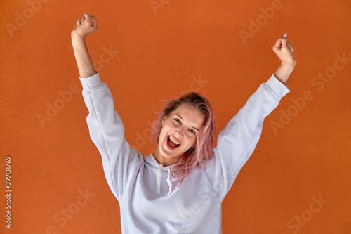 A beautiful happy young woman with pink hair holds her hands up and looks at the camera. Orange background. The concept of winning.