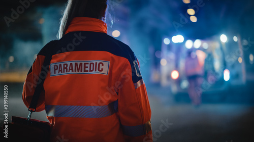 Female EMS Paramedic Proudly Standing With Her Back Turned to Camera in High Visibility Medical Orange Uniform with "Paramedic" Text Logo. Successful Emergency Medical Technician or Doctor.