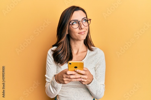 Young brunette woman using smartphone over yellow background smiling looking to the side and staring away thinking.