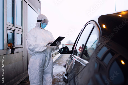 Medical heath care worker in protective white suit controlling passengers and PCR test at border crossing due to global corona virus pandemic.