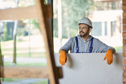 Handsome young male builder in hard hat looking positive, holding drywall while working at construction site