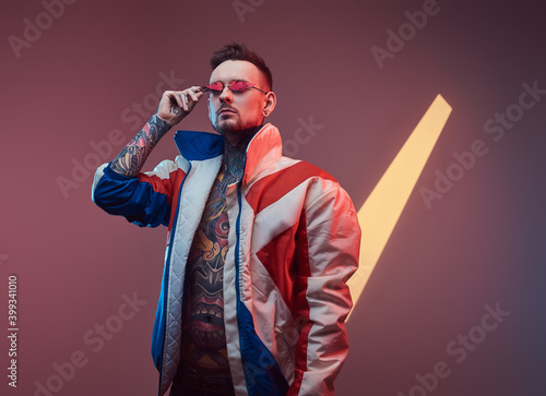 Fashionable and bearded hipster person with sunglasses and tattooed nude body dressed in warm winter coat poses in colourful background with serious face.