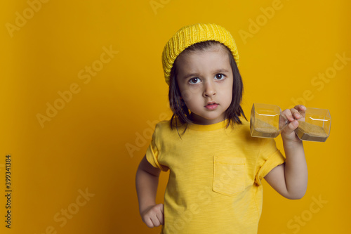 boy child in a yellow t shirt and a knitted hat on the background in the Studio holding an hourglass