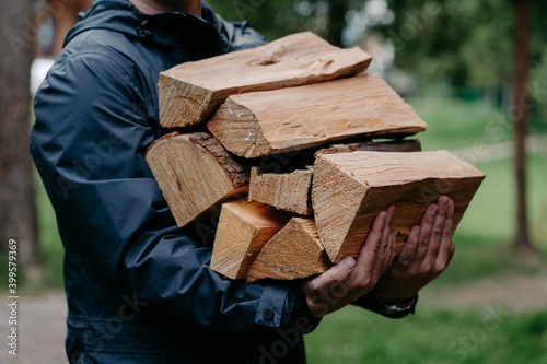 Cropped shot of faceless man in black jacket carries pile of firewood poses against blurred forest background.