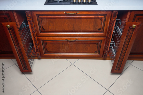 Pull out spice rack cabinet filler pantry. Two vertical spices drawers shelf under countertop with wooden panel and hob. Classic style kitchen with golden elements cherry alder wood cabinetry.