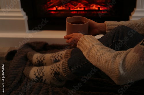 Woman in warm socks with cup of hot drink resting near fireplace at home, closeup