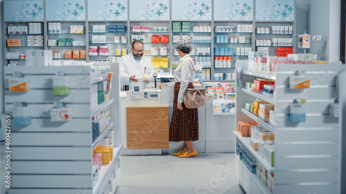 Pharmacy Drugstore Checkout Cashier Counter: Senior Woman Buying Prescription Medicine, Drugs, Vitamins and Talks to Beautiful Pharmacist Cashier, Asking Recomendations. Store with Health Care Goods