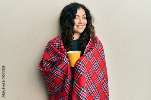 Young brunette woman with curly hair wrapped in a red warm red blanket drinking coffee winking looking at the camera with sexy expression, cheerful and happy face.