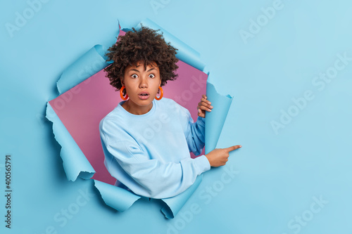 Shocked African American woman astonished by awesome advertisement indicates on right at blank space wears casual sweater poses through paper background in hole. People and promotion concept