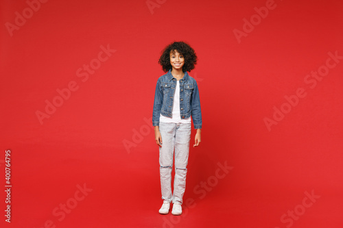 Full length of smiling little african american kid girl 12-13 years old in casual denim jacket looking camera isolated on bright red background children studio portrait. Childhood lifestyle concept.