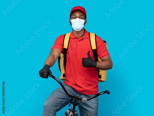 Black deliveryman in mask riding bicycle showing thumbs up