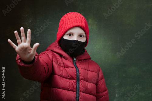 Boy with safety mask from coronavirus in winter jacket and red hat with raised hand in a gesture stop and protect Covid-19 outbreak around the Еarth
