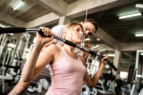 Cheerful young woman wearing pink sports bra while doing chin-up exercise with trainer man