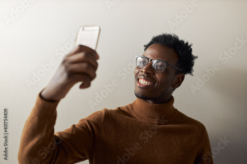 Cute cheerful young Afro American male holding cell phone using front camera for video conference chat, sharing good news, having excited facial expression. Black guy taking selfie on mobile
