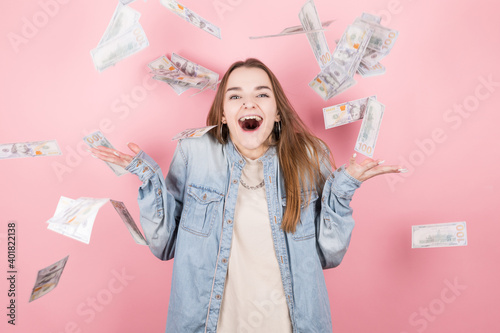 Portrait of pretty charming positive girl standing under shower from money having a lot of money in hands isolated on pink background
