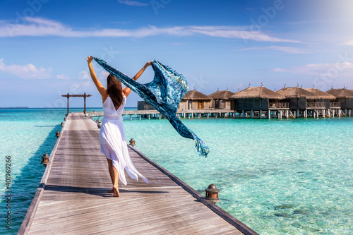 A beautiful tourist woman walks on a wooden pier over turquoise ocean in the Maldives and holds a waving scarf in her hands