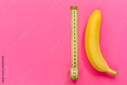 Yellow banana with measurement tape on pink background. Men penis size concept. Flat lay, top view, copy space.