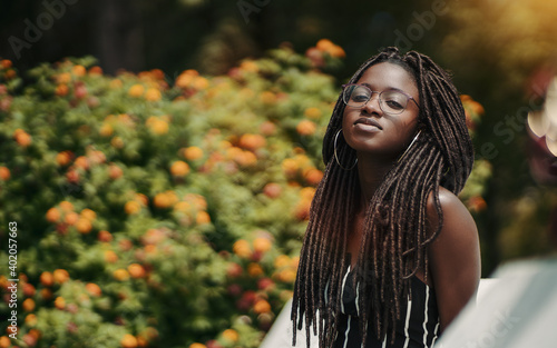 A ravishing young black female with dreadlocks and eyeglasses in the public park with a bush with flowers in the defocused background; a portrait of a dazzling African woman with dreads and in glasses