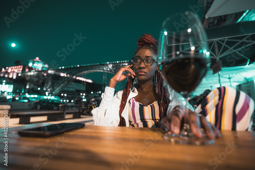 A pensive young ravishing black woman in eyeglasses and with braids is chilling in a night outdoor cafe with a glass of red wine in the defocused foreground; an arc bridge in the background, full moon