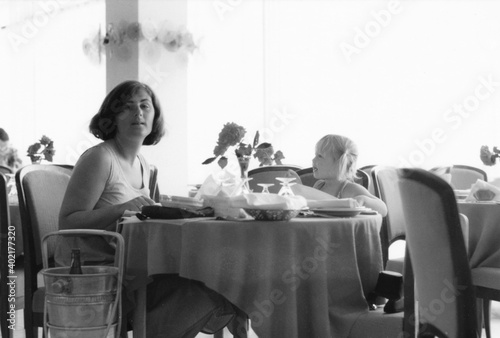 1977 vintage, seventies, retro monochrome image of a young mother and girl with blond hair sitting at a dining table in a restaurant of a hotel in Romania.