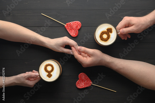 Top view Hands of couple in love holding lollipops and cups of coffee with symbols of venus and mars on milk foam on dark wooden table. Concept romantic date on Valentine's day. Creative flat lay