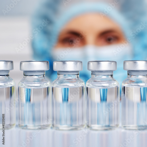 Female nurse in safety mask and cap looks at vials with liquid medicine on refrigerator shelf