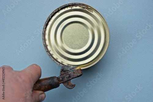 hand holds an old can opener that opens a can with a yellow lid on a gray table