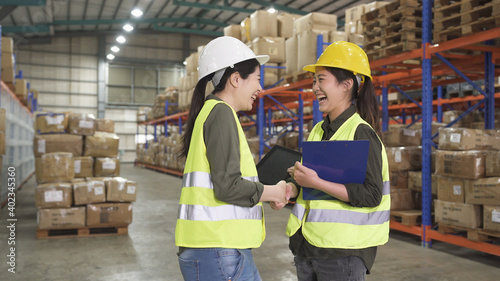 Warehouse operator teamwork. two laughing female storehouse operators shaking hand in large depot in company. best team partners women wear hard hat and safety vest doing handshaking and smiling.