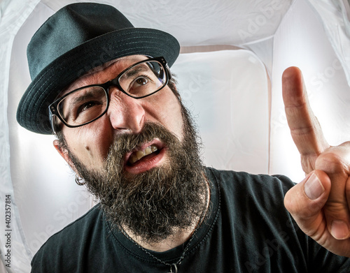 A bearded man with black glasses and hat showing finger and looking questioning