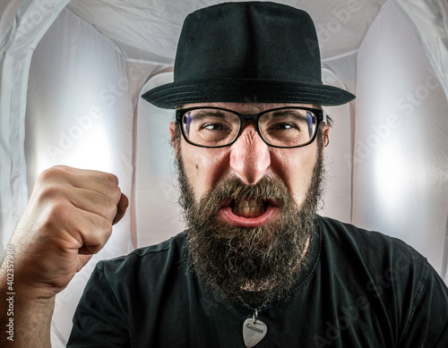 A bearded and angry looking man, looking like an artist or musician with black glasses and hat showing fist