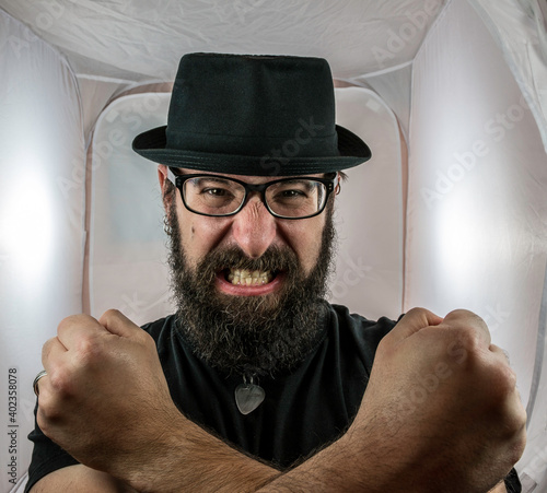 A bearded and angry looking man, looking like an artist or musician with black glasses and hat showing fist