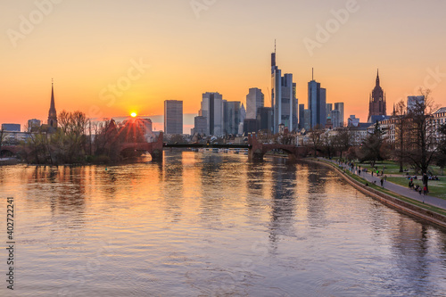 Sunset over the Frankfurt skyline. Skyscrapers, commercial buildings on the horizon. River Main with bridge and park on the bank. Reflections of the sun in the water